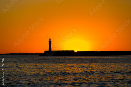 Sunset with the silhouette of the lighthouse in the background with flying birds and beautiful reflection on the water, calm afternoon on the beach in Almeria, Spain
