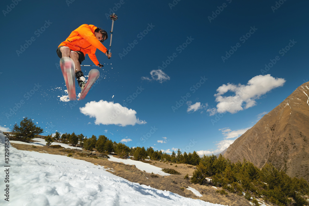 A young stylish man in sunglasses and a cap performs a trick in jumping with a kicker of snow against the blue sky and the sun on a sunny day.