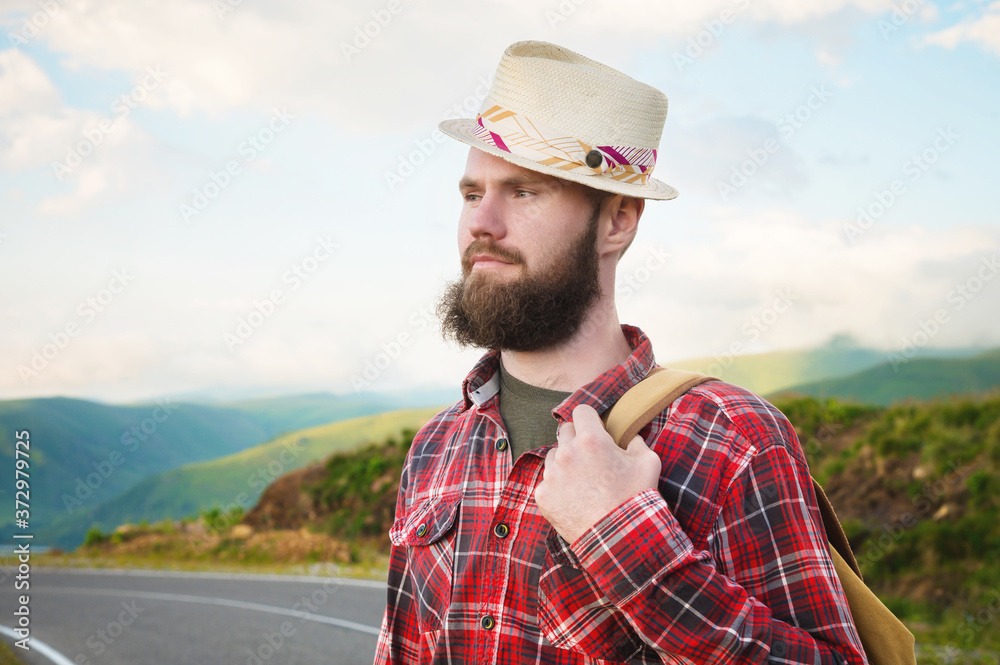 Portrait of a bearded happy smiling traveler hipster with a backpack in a plaid shirt and a hat next to an unknown car stands on the road at sunset in the mountains. Happy and confident travel concept