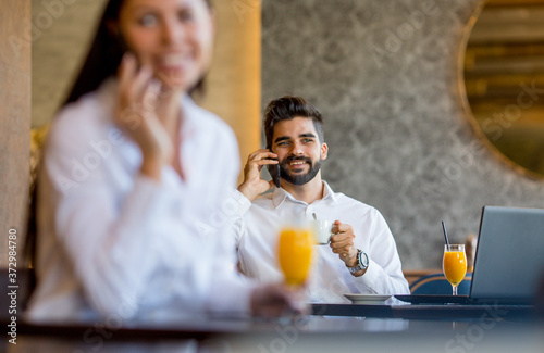A young woman is sitting at a table in a restaurant, talking on a mobile phone and drinking orange juice, behind her sits a young gentleman and talking on the phone