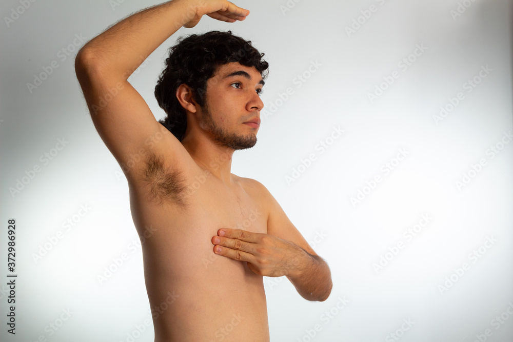 Obraz premium Young naked hispanic man with wavy hair and shaved beard, doing a checkup on his chest for signs of breast cancer