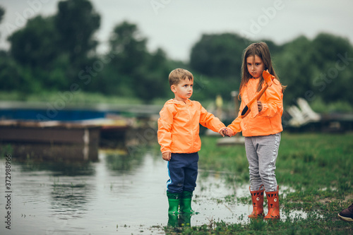 Brother and sister stand with boots in the river and hold hands on a gloomy day