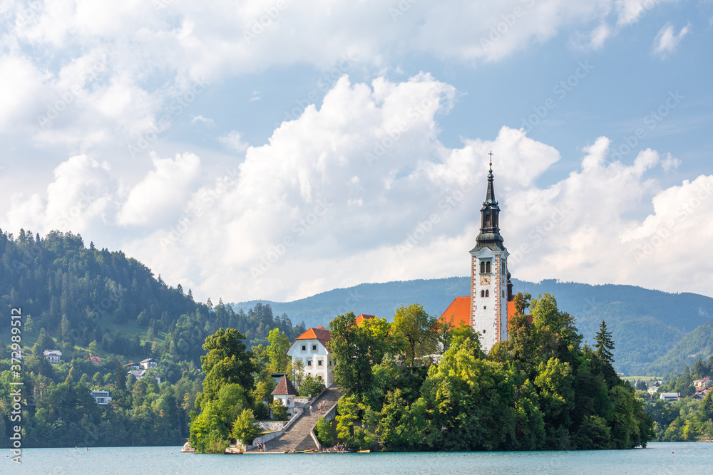 Close up of the iconic Slovenian island on the lake Bled surrounded by trees and vegetation, under a blue summer sky with puffy clouds