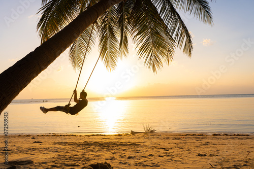 The guy enjoys the sunset riding on a swing on the ptropical beach. Silhouettes of a guy on a swing hanging on a palm tree, watching the sunset in the water.