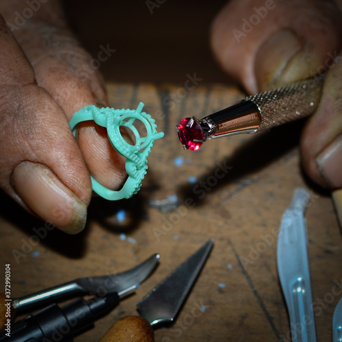 The male craftsman making jewelry is using a gemstone tool and a green wax holder to master the jewelry ring, and there are many tools for making jewelry in the jewelry industry on the wooden table.