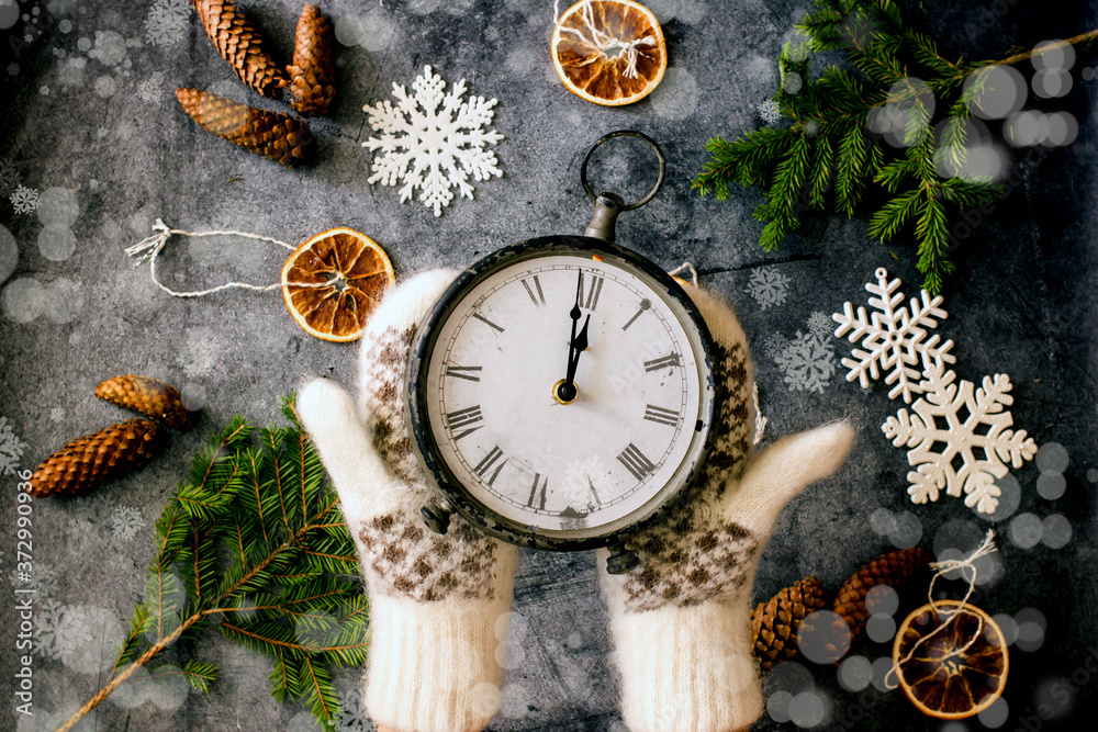 New Year's clock. Women's hands in mittens hold an antique clock showing midnight over a dark table with Christmas tree branches, cones, toys. Christmas background with clock and hand. Flat lay