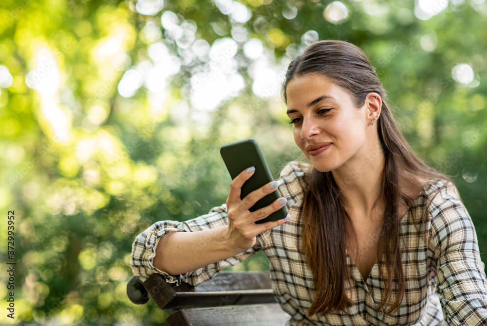 A young woman is sitting on a park bench and looking at her mobile phone A young woman is sitting on a park bench and looking at her mobile phone