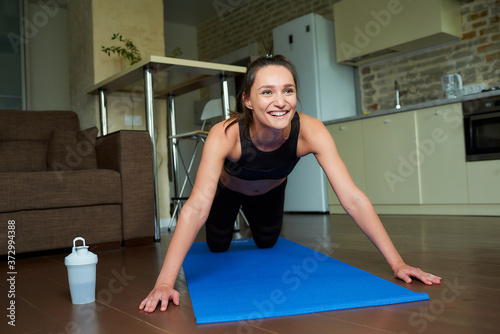 A smiling sporty girl in a black workout tight suit is laughing after doing push-ups on the blue yoga mat at home. A woman is relaxing after exercises for the chest at her apartment.
