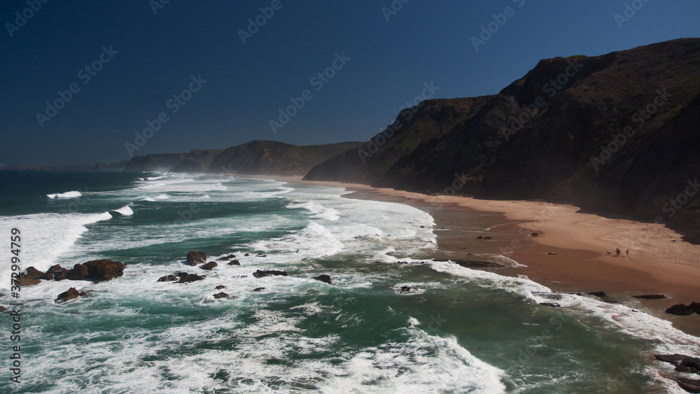 Top ocean coast view with sandy beach, black rocks, blue sky and huge waves