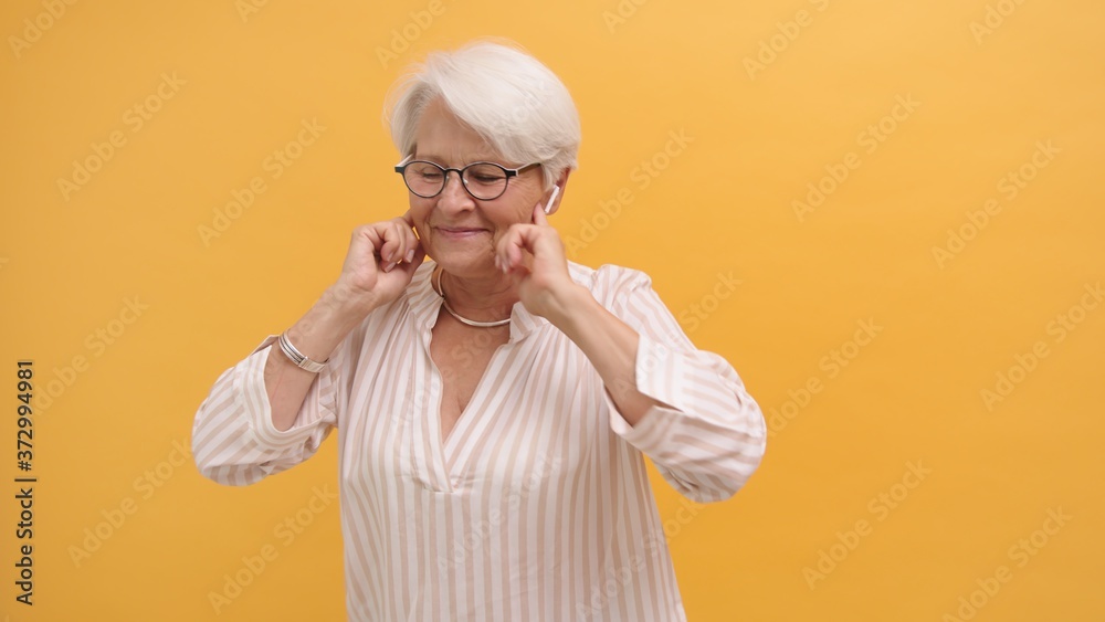 Old woman listening to the music with wireless headphones, isolated on orange background. High quality photo