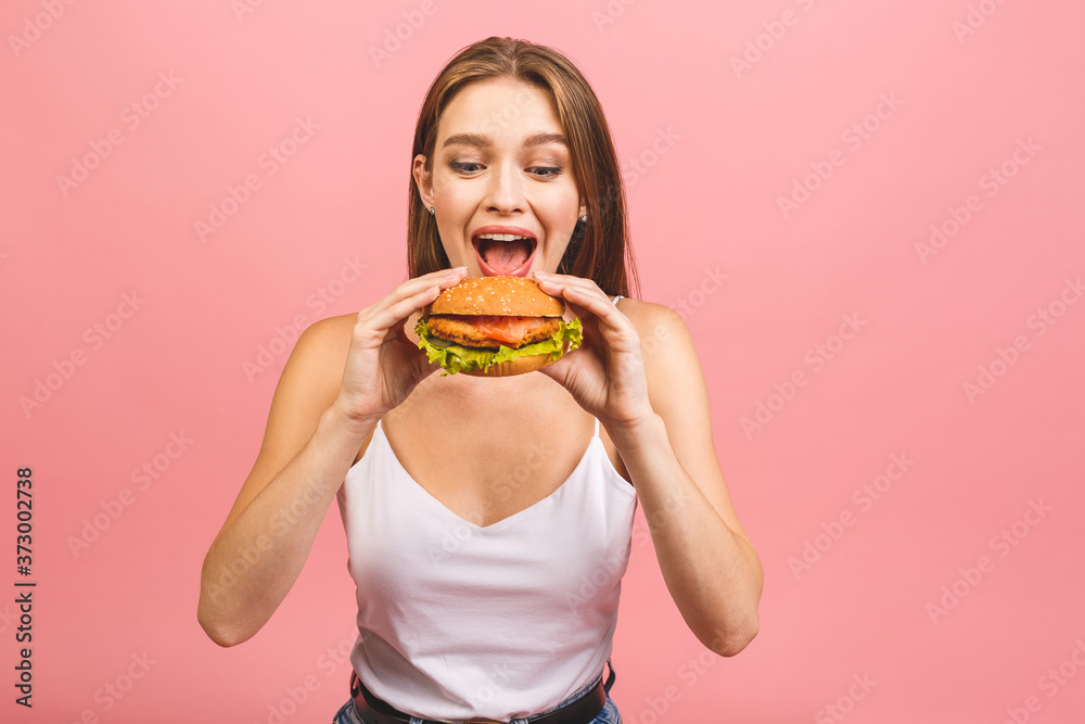 Portrait of young beautiful hungry woman eating burger. Isolated portrait of student with fast food over pink background. Diet concept. Portrait of young beautiful hungry woman eating burger. Isolated portrait of student with fast food over pink background. Diet concept.
