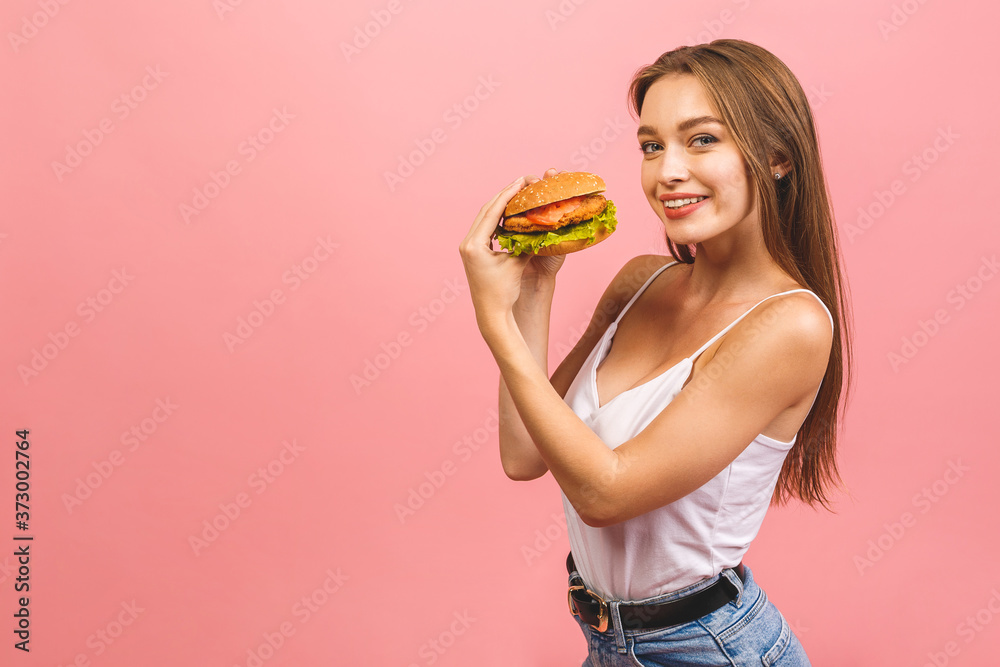 Portrait of young beautiful hungry woman eating burger. Isolated portrait of student with fast food over pink background. Diet concept. Portrait of young beautiful hungry woman eating burger. Isolated portrait of student with fast food over pink background. Diet concept.