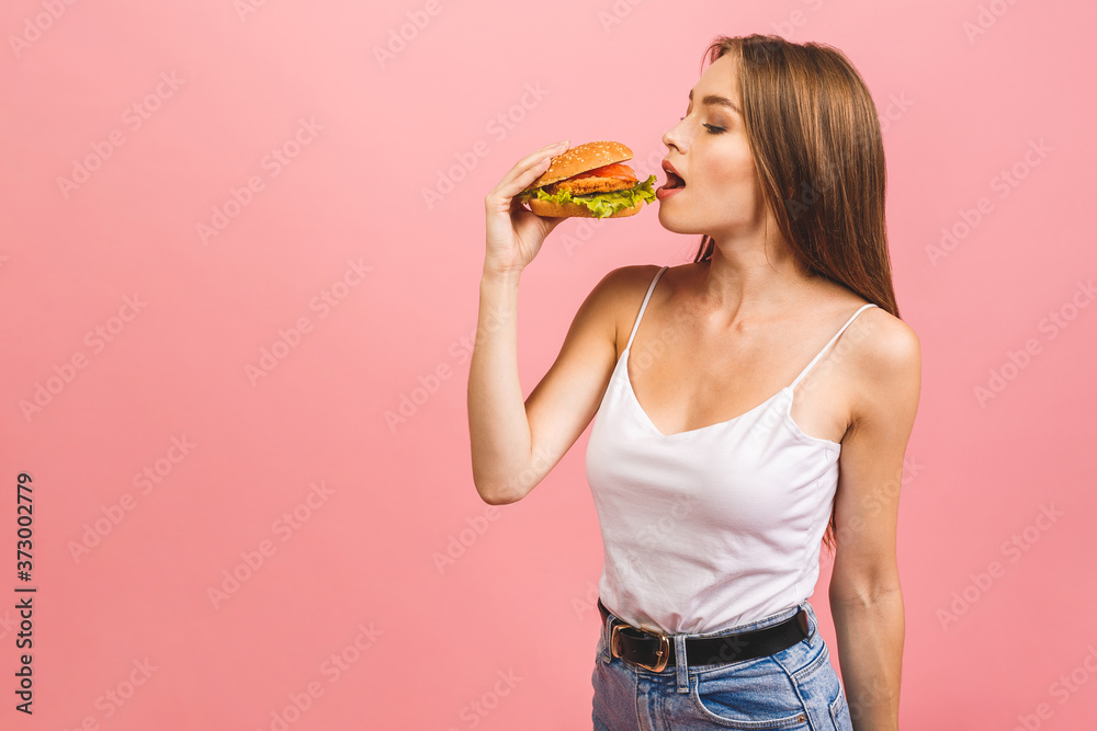 Portrait of young beautiful hungry woman eating burger. Isolated portrait of student with fast food over pink background. Diet concept. Portrait of young beautiful hungry woman eating burger. Isolated portrait of student with fast food over pink background. Diet concept.