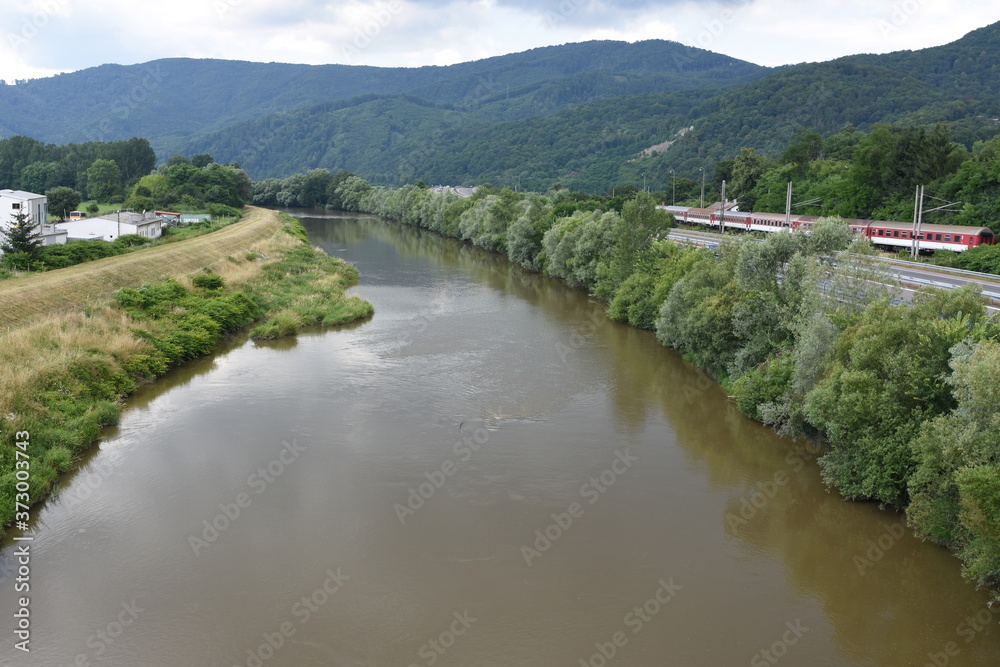 Fototapeta premium View from the bridge on Sedimentary water of river Hron in Slovakia near town Zarnovica. Banks of the river are covered by trees and bushes where is also visible electric train. 