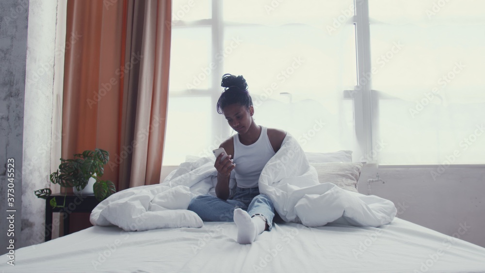 Pretty Young African American Woman uses phone while Relaxing in Bed