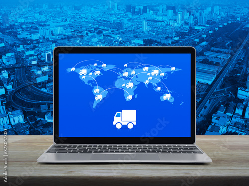 Delivery truck flat icon with connection line and world map on modern laptop computer screen on wooden table over city tower, street, expressway and skyscraper, Business transportation online concept