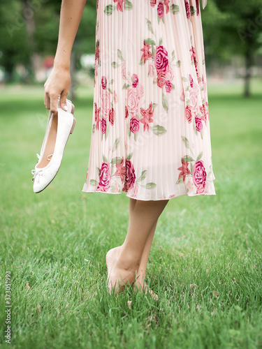 Young pretty woman in summer dress walking on meadow on green grass. A young girl holds white ballet shoes in her hands.