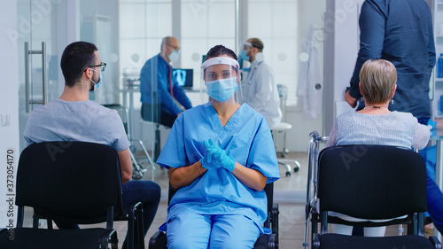 Medical nurse with face mask and visor against coronavirus wearing blue uniform in hospital waiting area. Doctor with senior man in examination room. Disabled mature woman in wheelchair.