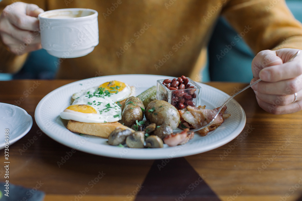 Traditional English breakfast with fried eggs, sausages, beans, mushrooms, and bacon on on a plate and cup of coffee on wooden table.