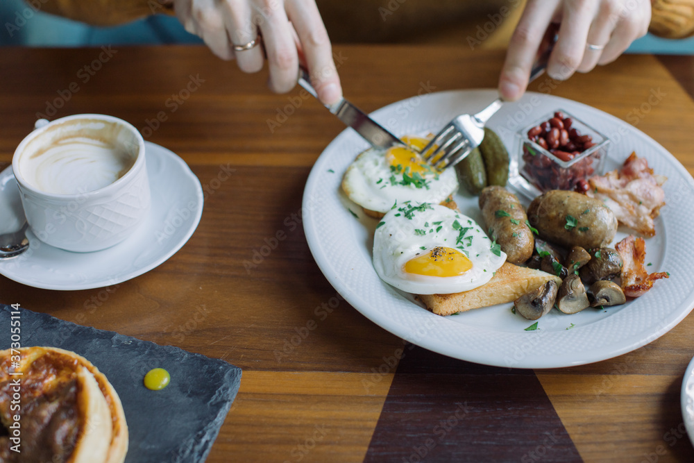 Close up of man's hands holding fork and knife while eating traditional English breakfast with fried eggs, sausages, beans, mushrooms, bacon.