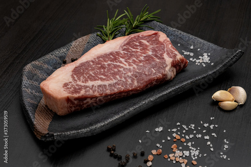 Raw of Black Angus Stiploin for cooking steaks with herbal ingredients garlic, rosemary, black pepper, himalayan pink salt in black tile plate on black wooden.Homemade cooking for relax your body.