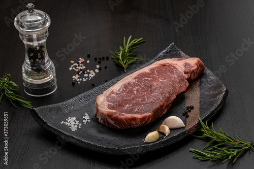 Raw of Black Angus Grain Fed for cooking steaks with herbal ingredients garlic, rosemary, black pepper, himalayan pink salt in black tile plate on black wooden.Homemade cooking for relax your body.