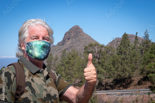 Serious senior man in surgical mask traveling in mountain landscape in Tenerife, gesturing ok sign - new normal life concept in summer vacation for an elderly people