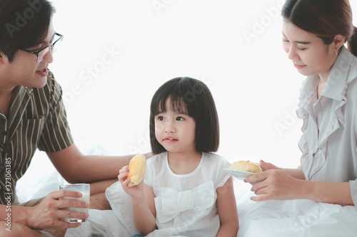 Happy family having fun in the bedroom. Father, mother, and daughter spending time together, girl eating bread and drinking milk, parents and kid having good memory together at home