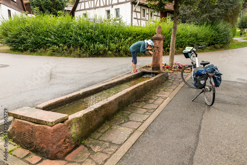 Fahrrad Fahrerin trinkt frisches Quellwasser aus einem Brunnen , willkommene Erfrischung auf der Fahrradtour. © Werner