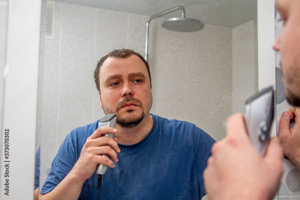A man shaves his beard and moustache with an electric razor at home in front of a mirror. Trimmer for cutting hair. A man shaves his beard and moustache with an electric razor at home in front of a mirror. Trimmer for cutting hair.