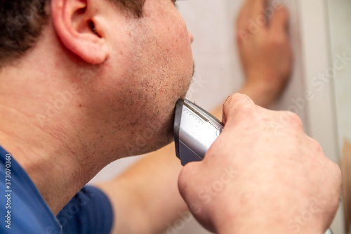 A man cuts his beard in the bathroom with a trimmer. Skin irritation from shaving. Short hairs fly from an electric razor.