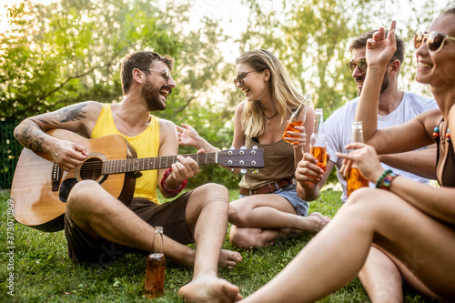 Two young couples sitting in the garden, enjoying and drinking beer on a sunny day while one of the men plays a guitar