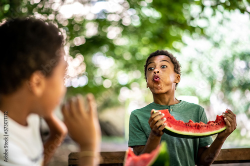 Two boys are sitting at a table eating a watermelon, one is spitting at the other with a watermelon seed photo