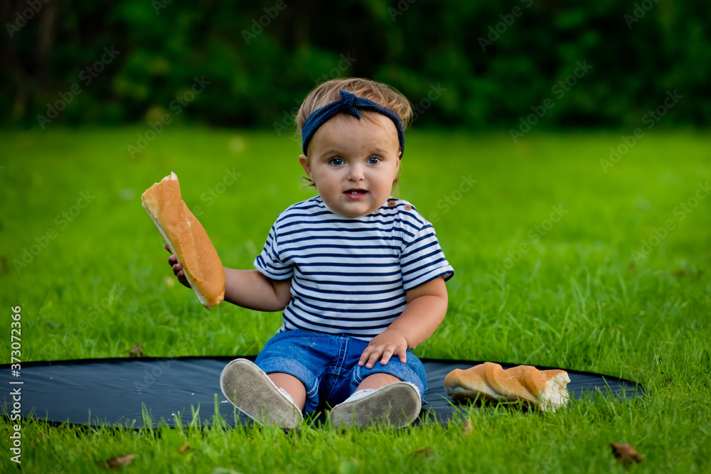 A little pretty baby girl sits on the lawn in the garden and holds a fresh baguette. A little pretty baby girl sits on the lawn in the garden and holds a fresh baguette.