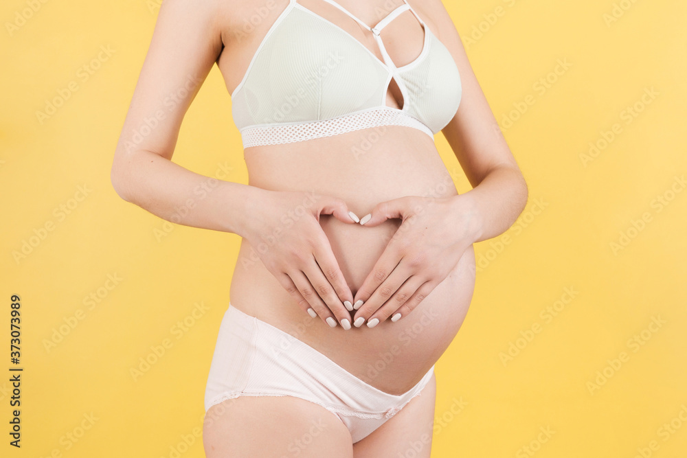 Close up of pregnant woman's belly with heart made of fingers at yellow background. Maternity concept. Love to the future baby. Copy space