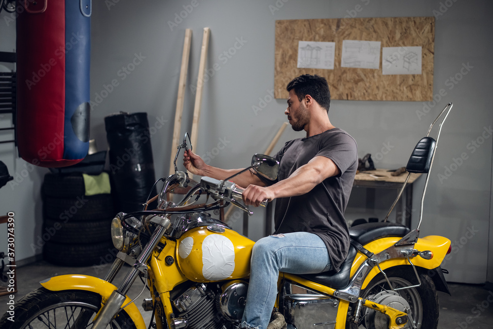 In the garage, a car mechanic sits on a yellow motorcycle that he has repaired. Pumped up stylish guy.