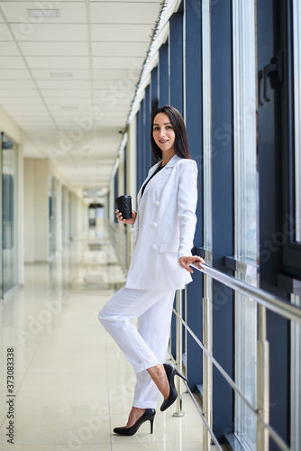 Young brunette woman, wearing white business suit, holding black paper coffee cup, standing in light office building near window. Businesswoman on lunch break. Corporate culture concept.