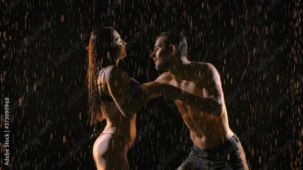 Passionate man and woman dance erotically and caress each other under streams of rain. Young naked bodies glisten with water droplets in studio light. Black background. Close up.