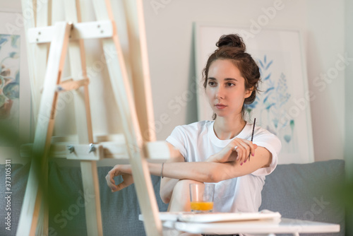 beautiful woman artist looks at a painting, in summer at home on couch, a brush in his hand, a table with paints and juice, a wooden easel background, a painting wall Woman's look. Free space.