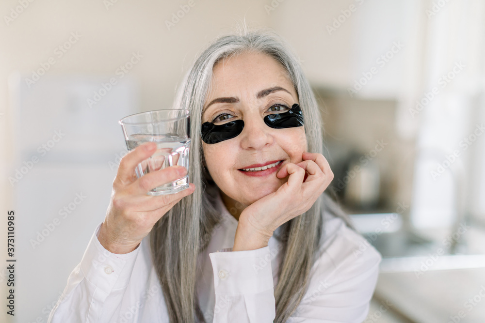 Elegant retired woman with long gray hair doing cosmetics procedures, posing to camera with black eye patches under eyes and glass with water. Beauty treatment at home kitchen interior Elegant retired woman with long gray hair doing cosmetics procedures, posing to camera with black eye patches under eyes and glass with water. Beauty treatment at home kitchen interior