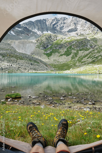 POV selfie shot of mans feet in trekking boots against majestic landscape of mountains and lake. Extreme hiker in tourist tent. Trekking lifestyle. Domestic local hike, tourism, travel.