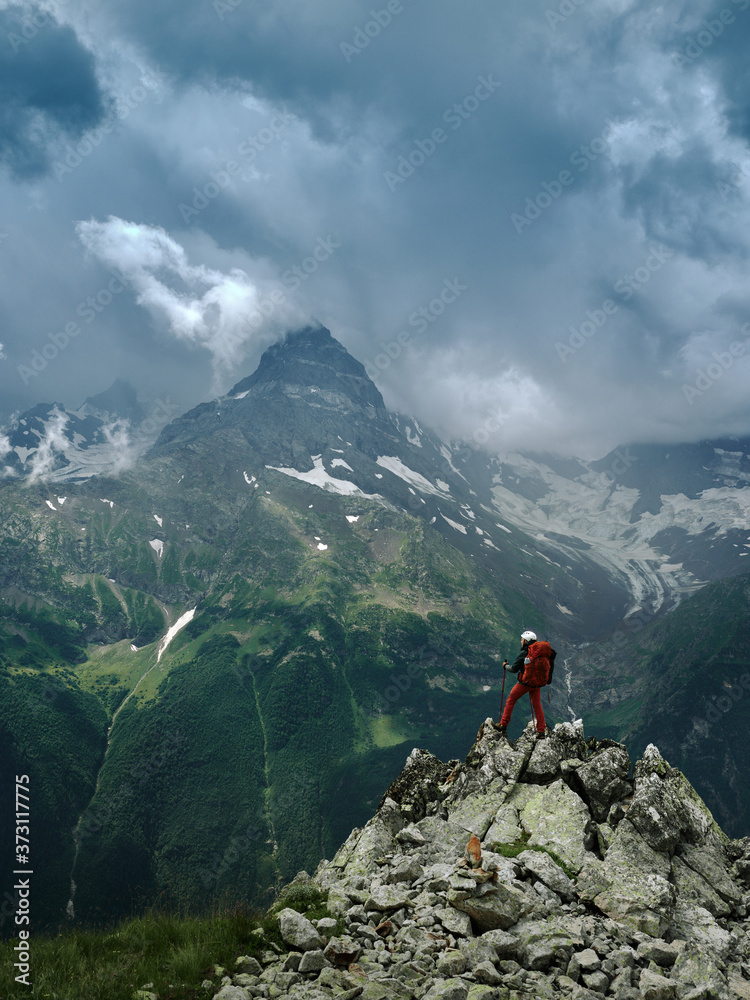 Alone hiker man with backpack and helmet against the gloomy mountain peak landscape with thunder cloudy sky, rocky ranges and peaks with glaciers. Domestic travel and trekking. Local tourism.