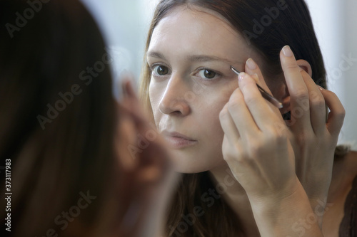 Young caucasian woman plucking her eyebrows in front of the mirror.