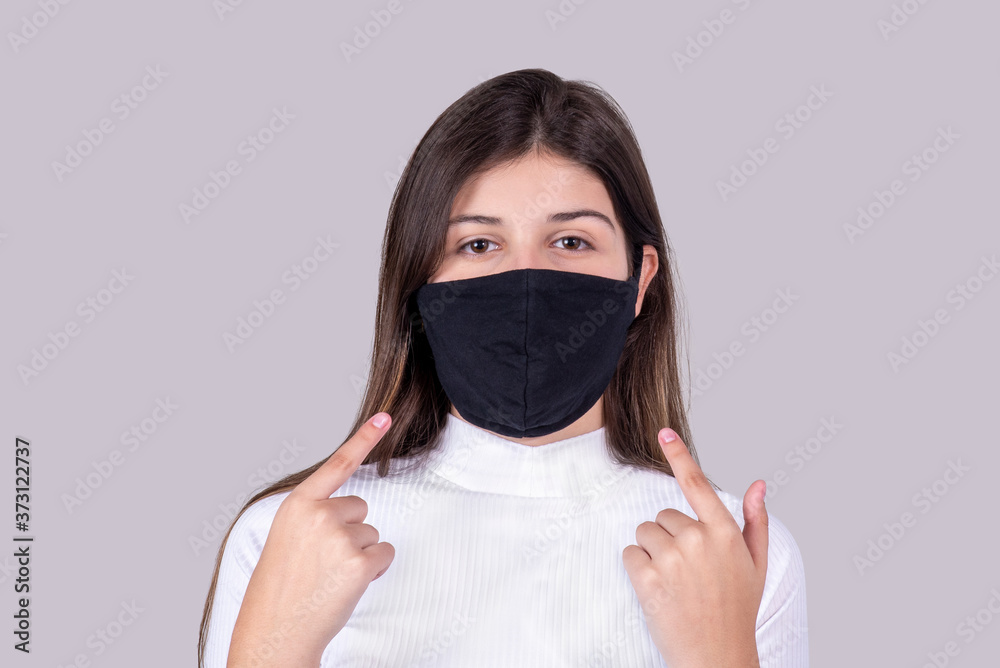 Young girl wearing a face mask looking at camera points her fingers to the mask. Isolated on gray background. Young girl wearing a face mask looking at camera points her fingers to the mask. Isolated on gray background.