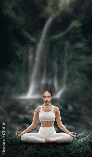 Beautiful young relaxed girl is engaged in yoga in the lotus position on the background of a waterfall in a tropical forest.