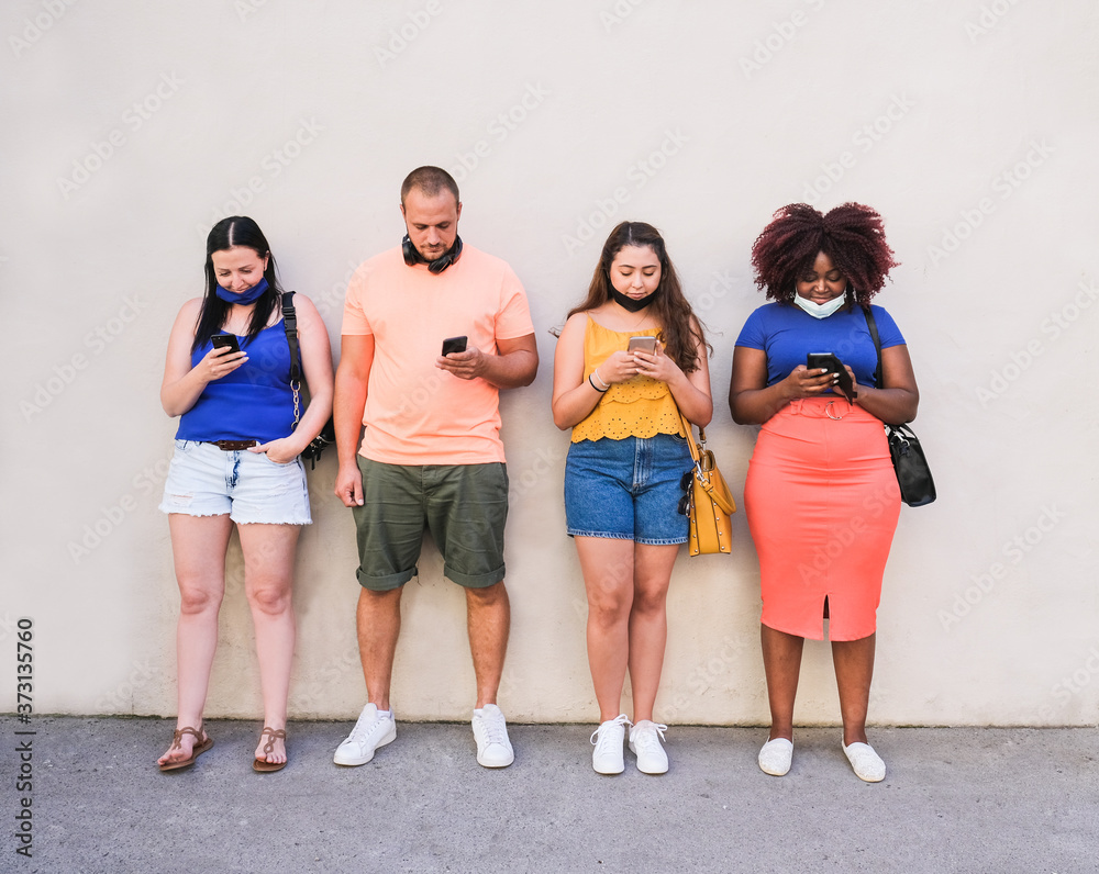 Obraz premium Young multiracial people standing in a line and looking at their mobile phones while wearing safety face mask - Coronavirus lifestyle