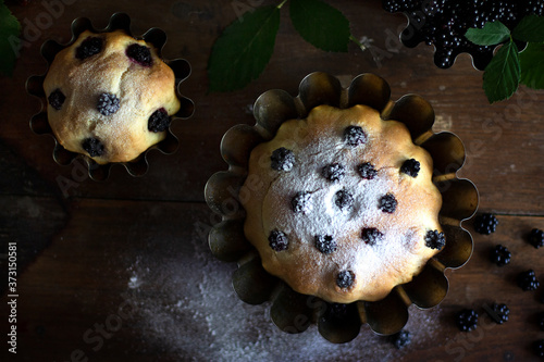 Delicious mouth-watering pastries on a vintage brown table with green BlackBerry branches. An open mailbox with a creative solution. Berry cupcake sprinkled with powdered sugar. Dark mood. Top view