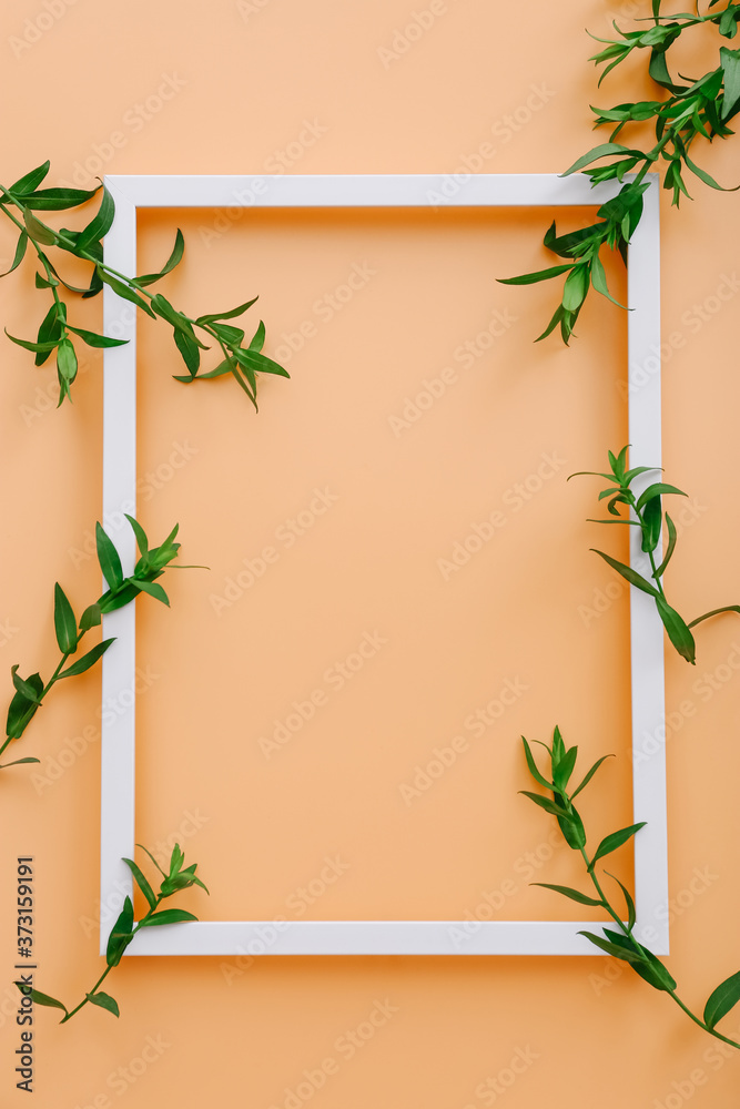 Empty picture frame with green leaves on peach color background, top view
