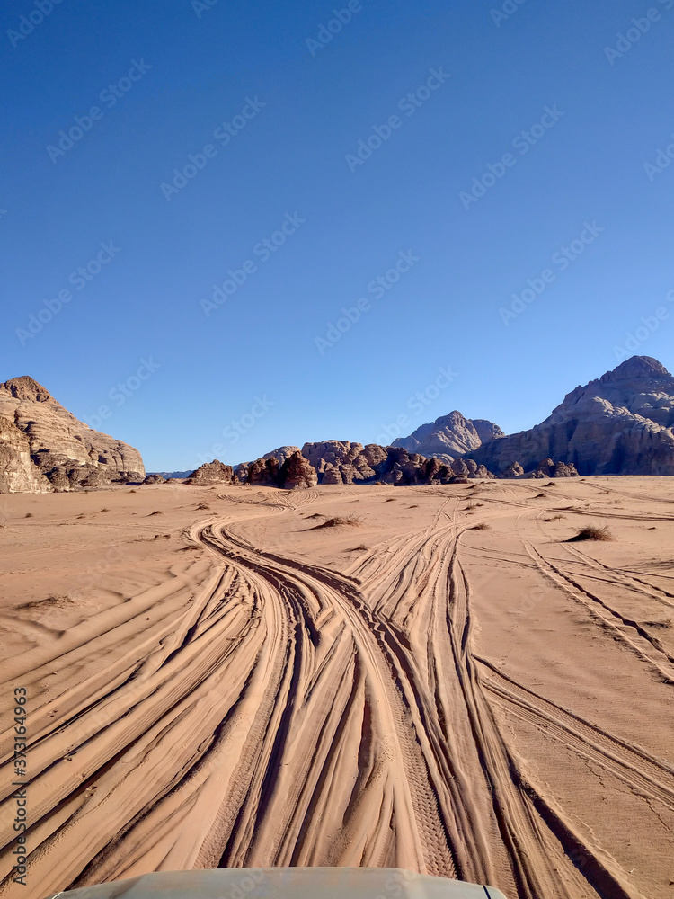 A car drives on a beaten track in the sands of the Wadi Rum desert in Jordan. The pickup truck hood is visible at the bottom of the photo. Clear blue sky. Theme of vacation in Jordan.