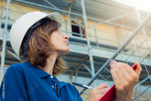 Woman, female engineer, caucasian, age 40, wearing a safety white cap, working on a costruction site in a typical men's role. Gender gap symbol.